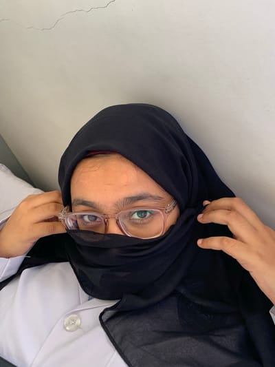The image shows a person wearing a black hijab and eyeglasses, with their face partially covered. They are wearing a white shirt or lab coat. The background is a plain white wall. The photo does not provide sufficient information to confirm the location is Picha Ya Ndege, Tanzania.