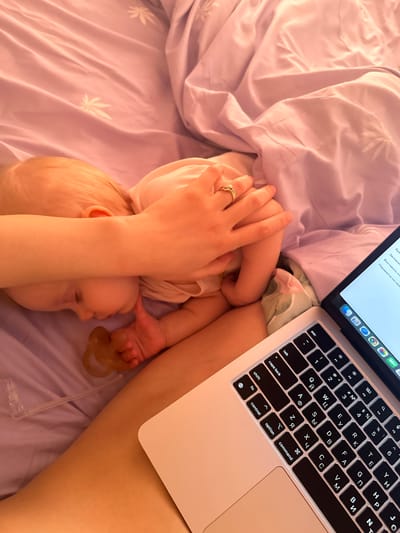 A top-down shot depicts a baby lying on a lilac-colored bedsheet with white leaf patterns, positioned next to an open laptop with a Cyrillic keyboard. The laptop's screen shows text in Russian. A person's hand, adorned with a ring, rests on the baby. A pacifier lies nearby on the bed. The shot appears to be taken indoors, possibly in Pokrov, Russia.