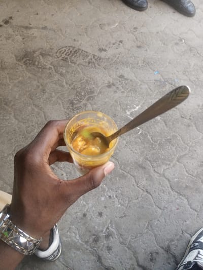 This image captures a downward-angled view of a dark-skinned hand holding a small, clear glass container. The hand, positioned slightly left of center, displays a silver-toned metallic bracelet with a structured, possibly studded or patterned, design on the wrist. Inside the glass is a thick, yellowish-orange substance containing visible solid chunks and lighter, possibly greenish, components. A silver-colored spoon, detailed with a chevron-like pattern on its handle, is partially submerged in the substance within the glass. The background consists of a grey, textured paved surface, consistent with interlocking pavers or concrete, showing signs of wear, discoloration, and a faint impression of a shoe sole. Portions of footwear from two other individuals are visible on the periphery: dark, possibly leather, shoes in the upper right quadrant, and a black, white, and grey athletic shoe with laces in the lower right quadrant. The scene is located in Majengo ward, Kenya.