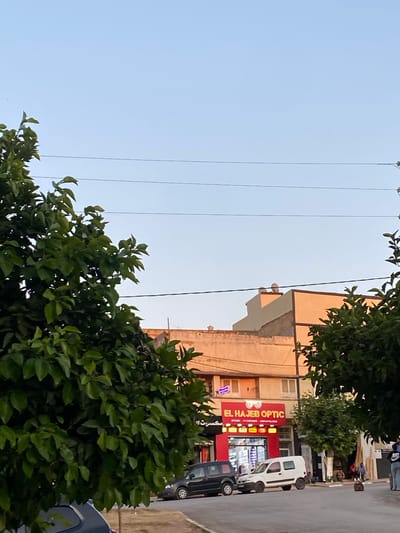 The image depicts a street scene in El Hajeb, Morocco. On the right side of the image, a shop with a red facade and illuminated signage reads "EL HAJEB OPTIC". The shop appears to be an optician's store. In front of the shop, a white van is parked, and a dark-colored car is also visible. Several people are seen near the shop, some sitting outside. The street is paved, and there are trees and utility poles lining the area. Buildings with orange-colored facades are present in the background, with clear blue sky overhead.