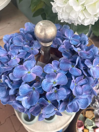 A close-up, overhead shot focuses on a cluster of artificial blue hydrangeas arranged around a silver sphere atop a wooden object. The hydrangeas have prominent, five-petal florets with a gradient of blue and purple hues. In the upper right background, a blurred white artificial hydrangea and green leaves are visible. The arrangement sits within a light-colored container, possibly ceramic, which is on a textured surface that appears to be a paved area. To the lower right, a blurred collection of small, colorful objects, possibly soaps or bath bombs, are partially visible. The scene is brightly lit, suggesting an indoor environment such as a shop or display. There is no discernible activity or interaction taking place.