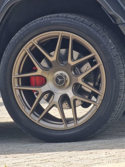The image shows a close-up of a car wheel. The wheel is a complex design with multiple spokes and a gold-colored finish. Behind the spokes, the car's braking system is visible, including a red brake caliper. The tire surrounding the wheel is black and appears to be high-performance. There are no people visible in the image. The setting is ambiguous and cannot be conclusively determined to be Witton Gilbert, United Kingdom, without additional visual or contextual information. The environment appears to be outdoors due to the available lighting.