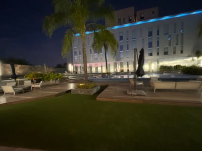 The image depicts the outdoor pool area of the Grand Fiesta Americana Querétaro hotel, located in the Juriquilla area of Santiago de Querétaro, Mexico, at night.

The scene is illuminated by artificial lights under a dark, clear sky. In the background, a modern, multi-story building, identified as the hotel, features a light-colored facade with numerous rectangular windows. A prominent blue LED light strip runs horizontally along the upper portion of the building.

The foreground and midground feature a meticulously maintained outdoor space. A large area of green turf, possibly artificial, occupies the immediate foreground. Beyond this, a network of light-colored wooden decking surrounds a central pool area. The pool is illuminated from within, displaying a light blue hue, and appears to have at least two distinct sections.

Several pieces of light-colored outdoor lounge furniture, including chaises and a sofa, are arranged on the wooden decking and an elevated concrete or stone platform. Multiple dark-colored patio umbrellas, some in a folded state, are positioned near the seating arrangements. Palm trees, one notably centered in the midground, and various potted plants are strategically placed throughout the area, contributing to the tropical ambiance. A low stone wall forms a boundary on the left side of the scene. No individuals are present in the image.