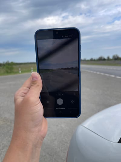 A person's left hand, holding a blue-cased smartphone, is angled towards the camera. The smartphone displays a camera interface, showing a road stretching into the distance under a cloudy sky. The background consists of a paved road with white markers and dry vegetation on either side, suggesting a rural or semi-rural environment. A portion of a white vehicle is visible in the lower right corner of the frame. The image appears to have been taken outdoors during the daytime. The location is specified as Moskovskoe, Russia.
