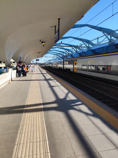The image shows a train platform at the Torino Airport & Caselle train station in Caselle Torinese, Italy. A long, silver and blue train with yellow accents is stopped at the platform. A group of people with luggage is gathered on the platform, waiting. The platform has textured tactile paving, gray tiles, and a yellow safety strip along the edge. Overhead, a modern architectural roof structure provides cover, casting shadows on the platform. The sky is clear and blue.