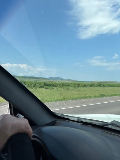 A first-person perspective shot from the driver's seat of a vehicle captures a landscape view through the windshield. The hand of the driver, adorned with a ring, is visible on the steering wheel. Outside the car, a vast expanse of green fields stretches towards rolling hills under a bright blue sky with scattered white clouds. A road with white lane markings is visible on the right side of the frame. The scene appears to be a rural or natural setting, possibly in or near Borodino, Russia, given the context.