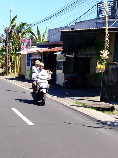 In Tabanan, Indonesia, two individuals, a driver and a passenger, travel on a white scooter down a paved road. The driver, wearing a white shirt and a white head covering, operates the scooter. The passenger, dressed in a white shirt and a tan cap, sits behind the driver. A shadow from the scooter and its occupants stretches across the road. To the left of the road, a vertical sign with the word "SALON" is visible, accompanied by a poster advertising an event. Behind the scooter, buildings with corrugated metal roofs and green facades line the street. Electrical wires crisscross overhead. A traditional Balinese decorative hanging, adorned with floral elements and yellow fabric, dangles from a structure on the right side of the scene. The ground beside the road features a narrow concrete walkway bordered by green grass and some debris. The sky above is a clear blue.