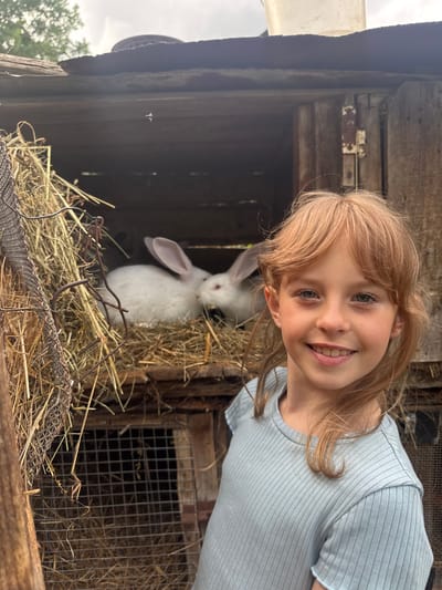 A young girl with blonde hair and blue eyes smiles at the camera, positioned in the foreground to the right of a wooden rabbit hutch. The hutch, appearing weathered and rustic, contains two white rabbits nestled in straw. One rabbit is more visible, facing towards the left side of the frame, while the other is partially obscured. The background depicts an overcast sky and hints of greenery, suggesting an outdoor rural or semi-rural setting. The overall scene evokes a sense of wholesome interaction between the child and the animals, within what appears to be a domestic or farm environment. The location in Малая Рукавицкая, Russia, is noted as context for the setting.