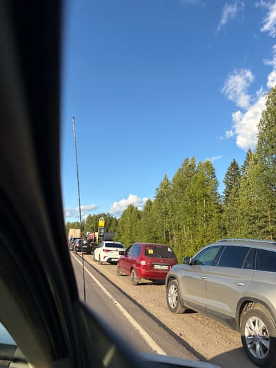 The image captures a traffic jam on a road outside the city of Kady, Russia. A convoy of cars is stopped on a two-lane asphalt road with a dirt shoulder. The vehicles are lined up in two lanes, with trees and foliage forming a dense forest along the right side of the road. In the background, a yellow sign with Russian lettering, likely indicating a gas station, is visible. The sky is a clear blue with scattered white clouds. The perspective suggests the photo was taken from inside a vehicle, looking forward. The overall scene depicts a common occurrence of traffic congestion, likely due to road conditions or construction, in a rural or suburban setting.