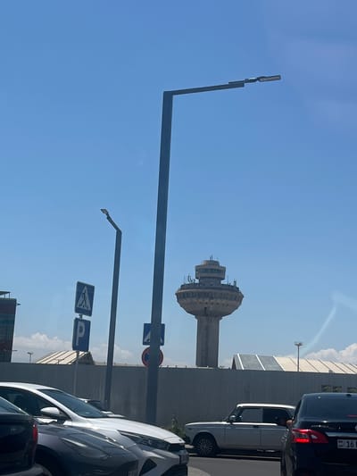 The image captures a street scene in Parakar, Armenia, on a bright, clear day. In the background, a distinctive Soviet-era control tower with a wide, tiered top stands against a vibrant blue sky. A grey, corrugated metal fence runs horizontally across the midground, partially obscuring the base of the tower and some modern building structures with angled roofs behind it.
In the foreground, several cars are lined up on a paved road, suggesting traffic. The leftmost vehicle is a dark-colored SUV, partially visible. Next to it is a white SUV or crossover. To the right of that is a white sedan, a classic model with chrome detailing. The rightmost vehicle is a black sedan, with a visible license plate reading "AM 16".
On the left side of the image, a tall, dark lamppost with an angled arm holding a light fixture rises vertically. Another lamppost, also tall and dark, is positioned to the right of the first and appears to have a light fixture. Several traffic signs are visible on poles: a blue square sign with a white pedestrian symbol, a blue rectangular sign with a white "P" for parking, and a blue square sign with a white symbol that is difficult to discern. A red and white circular sign with a red diagonal bar is also visible on a pole. The overall environment appears to be urban or on the outskirts of a city, with a mixture of infrastructure and vehicles.