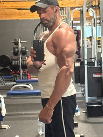 A muscular man is captured taking a mirror selfie in an indoor gym setting in Jemelle, Belgium. The man, seen from the chest up, exhibits a well-defined physique, with prominent muscles and visible veins on his right arm. He has a beard and is wearing a black baseball cap, a white sleeveless tank top featuring a dark, stylized emblem (possibly an 'M' or winged design), black track pants with a white stripe, a gold chain around his neck, and a gold bracelet on his right wrist. He holds a black iPhone in his right hand, positioned to take the reflection photograph.
The background of the image reveals various gym elements, including weight machines, racks loaded with numerous black weight plates, and what appears to be a clear water bottle on the floor. The lighting indicates an indoor environment, likely illuminated by artificial light. The man appears focused, seemingly showcasing his physique. No explicit time of day or weather conditions are discernible from the image. Aside from the stylized emblem on his shirt, no other specific text is clearly visible on any objects or equipment.