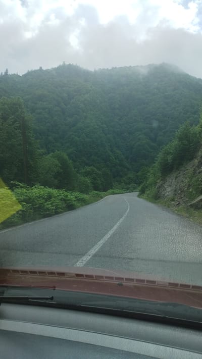 The image shows a view from inside a vehicle, likely a car, looking out onto a paved road. The road curves to the left. A white dividing line runs down the center. To the left of the road is a densely forested mountainside shrouded in mist or low-hanging clouds. The sky is overcast with grey clouds. Parts of the dashboard and windshield of the vehicle are visible in the foreground. Location information (Kostylivka, Ukraine) is provided, but not visually verifiable within the image.