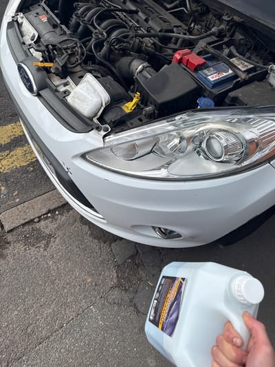 The image shows the front right side of a white Ford car with its hood open, revealing the engine bay. A person's hand is visible on the right, holding a translucent white bottle of screenwash, positioned as if to pour liquid into the car. This scene takes place outdoors on an asphalt surface, suggested to be in Boston, United Kingdom.

Inside the engine bay, a blue BOSCH car battery is prominent, along with various hoses, pipes, and reservoirs, one capped in blue. The car's front right headlight assembly, with its clear lens and reflective internal components, is clearly visible. The white bumper beneath the headlight shows signs of dirt and minor paint wear. The Ford logo is visible on the lower front grille.

The screenwash bottle has a label that reads "Ready to use SCREENWASH ALL SEASONS" in bold text, with additional smaller text and symbols. The liquid inside the bottle appears light blue.

The car is parked on an asphalt or tarmac ground, with yellow road markings (double lines) and a concrete curb visible, suggesting a roadside or parking lot setting. The bright, even lighting indicates it is daytime. The overall activity depicted is car maintenance, specifically the act of adding or preparing to add screenwash fluid to the vehicle.
