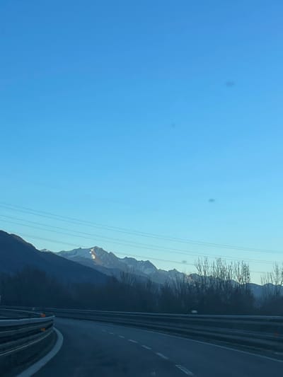 The image captures a clear, sunny day in a mountainous region, likely the Aosta Valley in Italy, viewed from a moving vehicle. The original image is oriented vertically, but depicts a horizontal landscape.
In the foreground on the right side, a multi-lane highway or road with metal guardrails is visible, suggesting travel. The road appears wet or reflective in parts. Beyond the road, dark, forested hillsides rise, leading to imposing mountains in the mid-ground and background. Many of the mountain peaks are partially covered in snow, with some catching the bright sunlight, creating a golden glow on their surfaces. Bare trees or branches are visible along the upper edge of the initial forested area, silhouetted against the bright sky.
The vast left side of the frame is dominated by a clear, vibrant blue sky, devoid of clouds. Several thin power lines stretch horizontally across the sky. Small, circular dust spots or lens flares are noticeable in the upper left portion of the blue sky. The scene conveys a sense of peaceful travel through scenic alpine landscapes. No people or visible text are present in the image. The time of day appears to be morning or late afternoon, given the angle of the sun illuminating the mountain peaks.