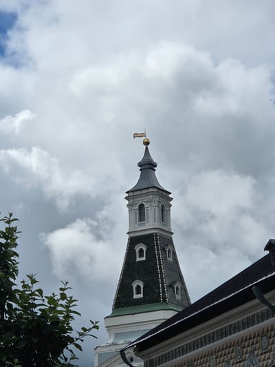 This media file depicts a low-angle view of a church spire against a cloudy sky. The spire has a grey, conical top adorned with architectural details and a golden orb at its peak, from which a small, golden banner hangs. The spire transitions to a section with grey and white decorative elements and arched windows, leading to a green-tiled roof with dormer windows. The scene is framed by the edge of a building with dark wood and ornate patterned trim on the right, and foliage from trees in the lower right corner. The sky is filled with dramatic, overcast clouds, suggesting either a coming storm or post-storm conditions. The lighting is diffused, indicating it is daytime, likely late morning or afternoon, without direct sunlight. No people are visible in the frame, and the focus is on the architectural structure and the sky. The image's perspective, looking upwards, emphasizes the grandeur and height of the spire. The context provided suggests the location is Sergiyev Posad, Russia, a place known for its significant religious architecture.