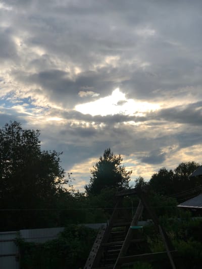The image depicts a backyard scene in Staroe Istomino, Russia, under a dramatic sky. Heavy, gray clouds dominate the upper portion of the frame, with patches of bright sunlight breaking through in the center, creating shafts of light. Below the sky, silhouetted trees and foliage fill the middle and lower sections of the image. To the left, a large, dark tree with dense leaves is prominent. In the center, a tall, thin evergreen tree stands out against the lighter sky. A wooden swing set with a green ring is visible in the foreground, partially obscured by the trees. A light-colored fence runs horizontally across the lower part of the frame, with overgrown greenery on top of it. A dark roof, possibly of a house, with solar panels is visible in the upper right corner of the image. The overall impression is one of a rustic outdoor setting during a cloudy, possibly stormy, weather event.