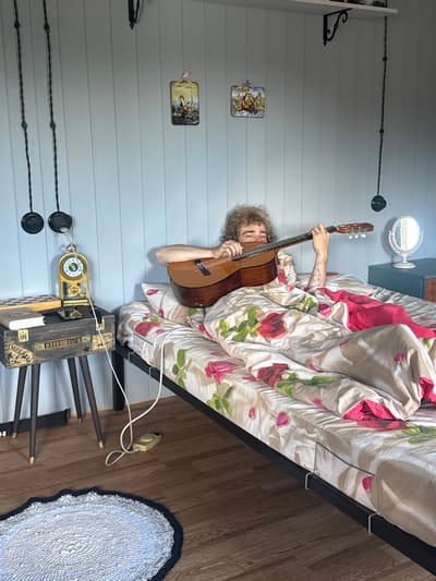 A person with curly blond hair is lying in a bed and playing a guitar. The person is covered by a blanket with a floral pattern of roses and petals. The bed frame is black metal. To the left of the bed is a nightstand with a vintage-style suitcase on top. On the suitcase are a book and a chessboard. A decorative clock is also on the suitcase, hanging from a wire. On the floor to the left of the nightstand is a round rug with a dark border. The walls are light blue and paneled. There are two small decorative pictures on the wall above the nightstand, and two black decorative objects hanging from wires. To the right of the bed is a dark teal dresser with a mirror on top. The scene appears to be in a bedroom. The image is not located in Desnogorsk, Russia.