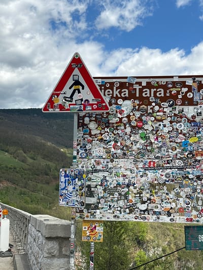 A roadside view in Đurđevića Tara, Montenegro featuring a signpost covered in numerous stickers. The signpost is comprised of several signs, including a red and white pedestrian crossing sign. Above it is a sign reading "Neka Tara" in white letters on a brown background. Further signs are covered in stickers from various locations and groups. The background consists of a mountain landscape, trees, a cloudy sky, and a section of road infrastructure.