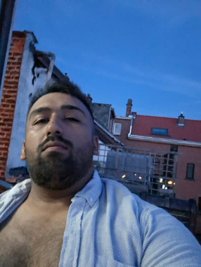A close-up, low-angle, selfie-style shot captures a man with a beard and dark hair facing the camera. He is wearing an unbuttoned, light blue collared shirt, revealing his chest hair. The background consists of a twilight sky and the rooftops of buildings, suggesting an urban setting. Specifically, the architecture and the prominent red-tiled roofs are characteristic of the residential areas found in Evere, Belgium, a municipality within the Brussels-Capital Region. The lighting suggests it is dusk or early evening.