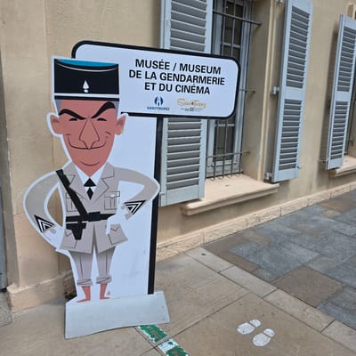 This image shows an outdoor sign for the "Musée / Museum de la Gendarmerie et du Cinéma" in Saint-Tropez, France. The sign is a large, vertically oriented rectangle with a white background and a black border. The text is in French and English, with the French text larger and bolder. Below the text is a cartoon illustration of a gendarme (French police officer) in a stylized uniform. The gendarme has a cheerful expression and is depicted in a somewhat humorous, exaggerated style. The sign is positioned outdoors, leaning against a building with light-colored stucco walls and light blue shutters. The ground in the foreground is paved with stone tiles. There are also two white footprints painted on the stone pavement, suggesting a path or a directional cue. A green and white sticker with some illegible text is visible on the pavement. The image appears to be taken during daylight, likely in fair weather. The overall impression is of a tourist attraction in a charming French town.