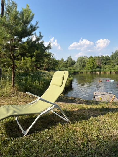 This image depicts a serene outdoor scene likely in Kamianka-Buzka, Ukraine, featuring a green lounge chair placed on a grassy area next to a calm body of water. The water is bordered by lush green trees and reeds, with a wooden pier extending into the water. Several inflatable tubes or swimming aids are visible in the water, suggesting recreational activity. The sky is a clear blue with scattered white clouds. The overall impression is one of a tranquil summer day in a natural setting.