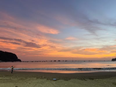 The image depicts a sunset over the bay of San Juan del Sur, Nicaragua. The sky is a gradient of purple and pink hues, with scattered clouds. The calm ocean reflects the colors of the sunset. Several boats are anchored in the bay, and a small hill or cliff is visible on the left side of the frame. On the sandy beach in the foreground, a person is standing with a child on their shoulders, pointing towards the water. The beach is dark and appears to be made of sand and small pebbles, with some debris visible. The overall scene is serene and picturesque, capturing the beauty of a tropical sunset.