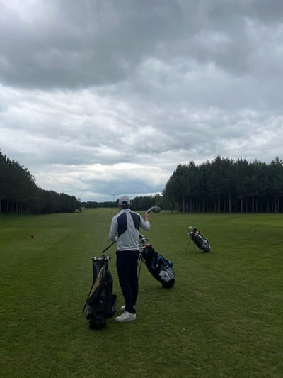 A man in a white golf shirt and a white hat is standing on a golf course in Skipsea, United Kingdom. He is holding a golf club in his right hand. He is looking at the golf club in his hand and about to throw it towards a golf bag. The bag is leaning against a golf club. There is another golf bag and golf clubs leaning against each other in the distance. The golf course is green and lush, and there is a line of trees on the right side of the image. The sky is cloudy and grey. In the background is a windmill, which is partially obscured by the clouds.