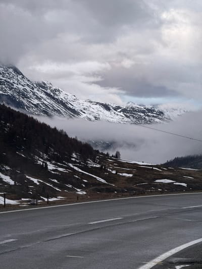 A majestic and rugged mountain landscape is depicted, likely in late winter or early spring, under heavy cloud cover. The image is oriented vertically, presenting a dramatic view with a paved road on the right and imposing mountains on the left.
The road, with distinct white lines marking its edge and center, stretches diagonally from the foreground into the distance, suggesting a mountain pass. To its left, a steep, rocky hillside is visible, partially covered in irregular patches of melting snow and sparse, dark vegetation. Further into the scene, a deep valley is indicated by a band of dark coniferous trees in shadow, partially obscured by mist or low clouds.
Dominating the left side of the frame is a grand, snow-covered mountain range, its jagged peaks and slopes blanketed in white. The intricate patterns of snow and rock are highlighted despite the diffused light. Swirling clouds and fog enshroud the middle and upper sections of these mountains, creating a sense of scale and mystique. A thin, prominent cable or power line stretches across the mid-air, cutting across the valley and mountains, hinting at infrastructure in this remote setting.
The overall atmosphere is one of dramatic natural beauty, solitude, and the raw power of the mountains. The weather appears cold and overcast, with the pervasive clouds adding to the somber yet awe-inspiring mood. The scene likely captures a view near Pontresina, Switzerland. There is no visible text.
