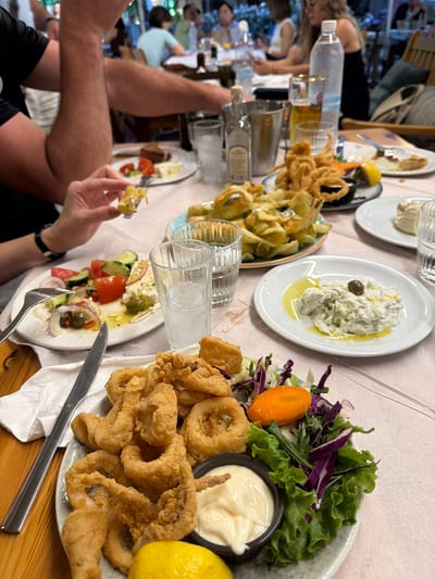 The image displays a detailed, slightly elevated perspective of a dining table in a restaurant located in Alexandroupolis, Greece, during what appears to be an evening setting. The foreground captures a meal in progress, with a person's dark-sleeved arm visible on the left and another individual's right hand, featuring painted fingernails, holding a fork with light-colored food over a white plate.
The table is draped with a light-colored tablecloth and is densely populated with various dishes and beverages. A prominent central dish is a large plate of golden-brown, breaded fried calamari rings and tentacles, accompanied by a lemon wedge and a side salad composed of green lettuce, shredded red cabbage, thin green vegetable strips, and a slice of orange carrot. Adjacent to the calamari is a small black bowl containing a white, creamy sauce. A silver knife rests on a crumpled white napkin beside this plate.
Other identifiable dishes include a white plate featuring Tzatziki, garnished with a single dark olive and olive oil; a partially consumed Greek salad (Horiatiki Salata) with sliced tomatoes, cucumbers, red onions, olives, and crumbled feta cheese, drizzled with olive oil; a plate filled with fried zucchini chips; and another white plate holding a beige, creamy dip or spread.
Beverages observed on the table include a tall glass with ice and a clear liquid, a second similar glass partly obscured, a tall glass of amber beer, a large plastic bottle of water, and a silver ice bucket containing a clear bottle, likely a local spirit.
The background is out of focus, revealing multiple blurred figures of other diners seated at various tables, illuminated by ambient lighting, indicative of a bustling and social restaurant environment. The overall scene suggests a typical Greek dining experience featuring a variety of traditional mezedes (appetizers) and seafood.