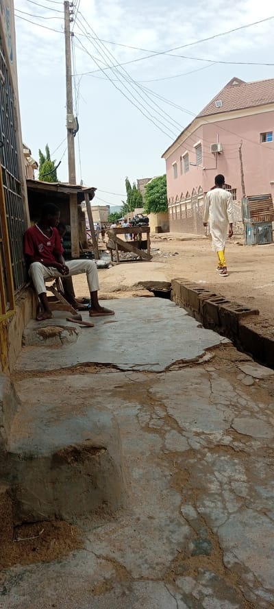 In Rijiyar Zaki, Nigeria, a dusty street scene unfolds. In the foreground, two young men are seated on a concrete ledge next to a makeshift wooden stall. The man on the left, wearing a red and white shirt and light-colored pants, faces away from the camera, his legs crossed. Beside him, a second individual, partially visible, wears similar attire. A wooden stool lies on the ground in front of them. The stall behind them is constructed from rough timber and covered with corrugated metal sheeting, with black bags stacked within.

Further down the street, a man in a white robe and bright yellow trousers walks away from the camera. He is heading towards a light pink building with arched windows and decorative elements along its facade, which appears to be a place of worship or a significant structure. In the middle ground, other individuals and market stalls are visible, suggesting a bustling commercial area. A utility pole with numerous wires stretches across the frame, indicating the presence of electricity infrastructure. The environment is characterized by unpaved roads, cracked concrete, and a generally dry, dusty atmosphere.