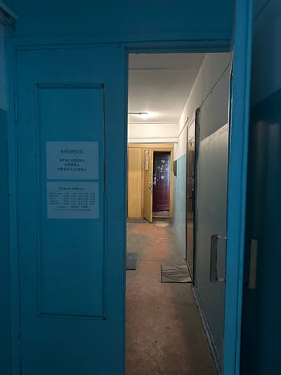 The image depicts an interior hallway with a door slightly ajar on the left, revealing a sign for a notary named Krasavina Irina Nikolaevna. The sign also lists the notary's working hours from Monday to Friday, 10:00 to 17:30 with a lunch break from 13:00 to 14:00, and on Saturday from 10:30 to 13:00. The hallway appears to be in a residential or administrative building, with a worn floor and blue-painted walls and doors. Further down the hallway, another door is visible, partially open, and appears to be made of dark wood with decorative elements. The scene is lit by artificial light, creating a somewhat dim and uninviting atmosphere. The image is taken from within a doorway, looking down the hallway towards the other end. There are no people visible in the image. Based on the provided information about the notary's location being Vilyuchinsk, Russia, this scene is likely within the city.