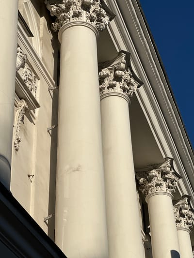 The image shows a close-up, low-angle view of the exterior of a building with classical architecture in Feklino, Russia. The building features large, white, fluted columns with ornate Corinthian capitals. The capitals are intricately carved with acanthus leaves and volutes. Above the columns, a white entablature with multiple moldings is visible, topped by a dark-colored roofline that slopes upwards against a clear, deep blue sky. Sunlight casts sharp shadows on the textured surfaces of the columns and architectural details, highlighting the dimensionality of the carvings and the building's facade. The wall behind the columns is a pale, textured cream color, with visible signs of age such as fine cracks and some faint discoloration. There are also some metal brackets attached to the wall. No people or animals are present in the image.