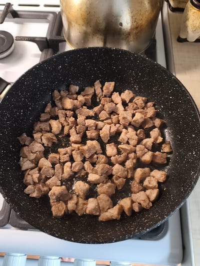 A black, speckled frying pan is filled with diced, browned meat that is sizzling in oil. The pan is situated on a white gas stovetop. In the background, a portion of a large, metallic pot is visible, also on the stovetop. The image appears to be taken from a slightly elevated angle, looking down into the pan. The overall scene depicts food being cooked in a kitchen environment.