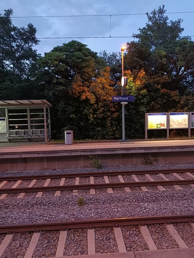 The image shows a train station platform with railway tracks in the foreground. The station sign reads "Harthaus," indicating the location. There is a covered waiting area with benches on the left, and information boards on the right. A lamppost with an illuminated lamp stands near the sign. Trees and foliage line the background, suggesting an outdoor environment. The sky is overcast, and the lighting indicates it is either early morning or late evening. The scene appears to be in Germering, Germany, as suggested by the name "Harthaus," which is a known locality in Germering. There are no people visible in the image.