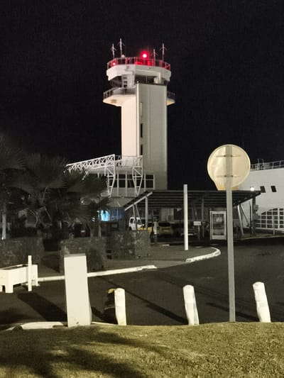 The image captures a night scene at an airport in Sainte-Marie, France. Dominating the frame is an air traffic control tower, its upper section illuminated by red warning lights. The tower is a multi-story structure with a white exterior, featuring a glassed-in control room at the top. In the foreground, a reflective circular sign on a pole stands to the right, casting a faint glow. To the left, palm trees and a stone wall create a tropical ambiance, incongruous with the typical French landscape. Behind the wall, a covered parking area shelters a yellow car and a white van. Other visible elements include white bollards, a grassy area, and a long, white building in the background. The scene appears deserted, with no people visible.