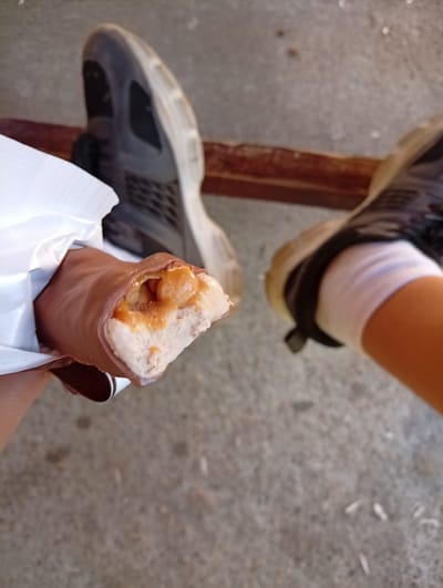 The image shows a close-up of a person's hand holding a partially eaten chocolate bar. The chocolate bar has a nougat filling and appears to be a "Snickers" bar. The person's feet, wearing athletic shoes, are visible in the background, suggesting they are outdoors. The ground is concrete, and there is a wooden plank or beam visible. The lighting indicates it is daytime. The scene appears to be casual, with the person taking a break to eat a snack. There is no visible text in the image.