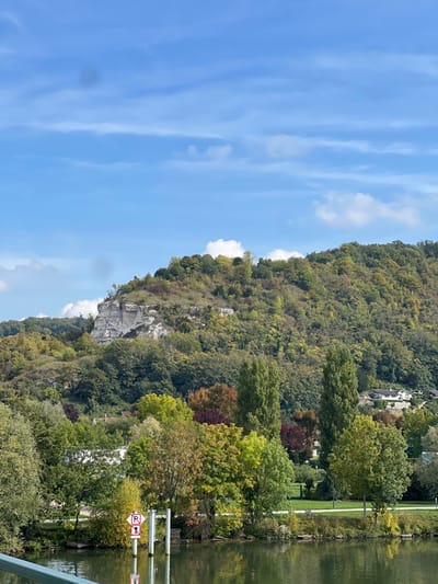 This image displays a landscape view of the Seine River in Vernon, France. In the foreground, the dark green river surface occupies the lower third of the frame, with a dark green bridge railing visible at the bottom left. Two white cylindrical poles extend from the water near the left bank; one features a circular red and white sign indicating "No Mooring" or "No Anchoring." The opposing riverbank presents a narrow strip of green land and diverse deciduous trees, exhibiting autumnal colors including green, yellow, orange, and red foliage. A prominent, densely forested hillside rises in the background, featuring a distinct, irregular white chalk cliff face exposed on its upper left slope. Several white residential buildings are partially visible amidst the trees on the hillside. The sky above is clear blue with scattered white stratocumulus clouds. No individuals are present in the scene.