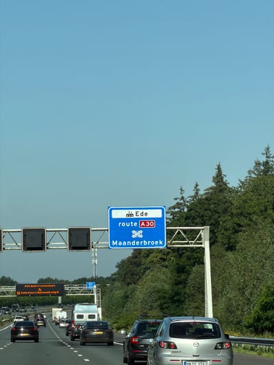 The image shows a highway scene with several cars and signs. A large blue overhead sign indicates "Ede" and "route A30 Maanderbroek." To the left, another electronic sign displays "A30 dicht ri Amersfoort" and "Amersfoort/Barneveld volg 5." Several vehicles are visible on the multi-lane highway, including sedans, a van, and a camper. The environment is characterized by a clear blue sky and lush green trees lining the side of the road. The overall impression is a typical highway landscape in the Netherlands, likely near Bennekom, given the mention of Ede and Amersfoort.