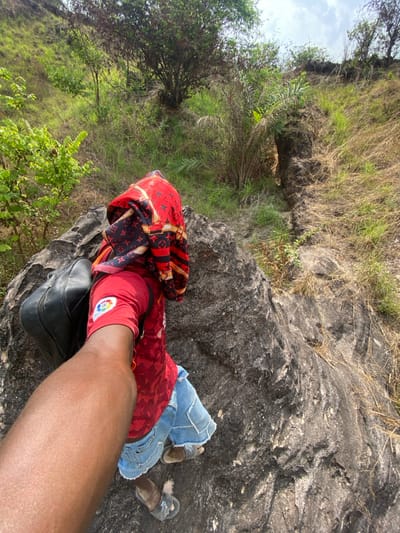 The image depicts an outdoor scene in Amoso, Nigeria, viewed from a high-angle perspective looking down a rocky slope. In the immediate foreground, a dark-skinned forearm and elbow are visible extending from the left, holding a black handbag. Below and slightly right, a person is positioned on a large, textured grey rock, facing away from the camera. This individual wears a red patterned shirt with a white and colored logo on the left sleeve, blue denim shorts with frayed hems, and dark-colored sandals. A red, black, and gold patterned fabric is draped over their upper back and the rock. A second black handbag rests on the rock near the person's left side. The surrounding environment consists of steep, uneven terrain characterized by large grey rock formations, patches of dry brown grass, green shrubs, and scattered trees. The sky, visible at the top, is light blue with sparse white cloud cover.