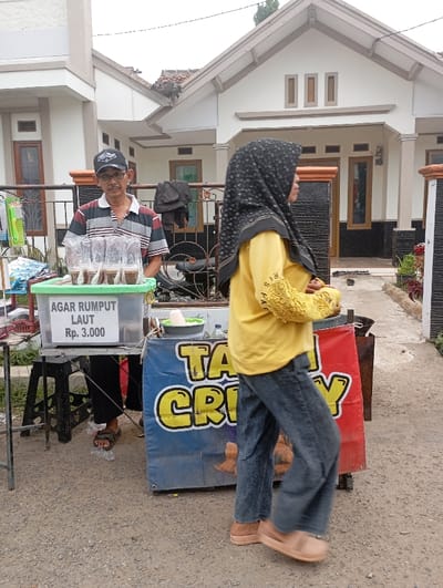 The image captures an outdoor scene in Cilampeni, Indonesia, featuring two individuals and street vendor stalls. On the left, a middle-aged man with a mustache, wearing a black baseball cap, glasses, and a striped shirt, stands behind a vendor setup. His stall includes a clear plastic container labeled "AGAR RUMPUT LAUT Rp. 3.000" (Seaweed Jelly Rp. 3,000), holding several cups of dark-colored contents, resting on a stand supported by two black stools. To the man's right, a woman is observed in mid-stride, walking from right to left. She is wearing a patterned black hijab, a long-sleeved yellow shirt, blue jeans, and light brown slip-on shoes; her face is turned in profile away from the camera. Adjacent to the seaweed jelly stall, a second vendor display features a blue and red banner with partially visible yellow text, "TA... CRISPY." The immediate ground is an unpaved, gravelly surface. In the background, two single-story residential houses with light-colored facades, dark roofs, and brown-framed windows are visible.