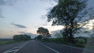 The image is a view from a vehicle driving on a road in Sokolovo, Bulgaria. The road is asphalt with white and yellow line markings. Trees line the sides of the road. The sky is overcast with clouds and the time appears to be dusk or dawn. The trees in the foreground are blurred indicating movement.