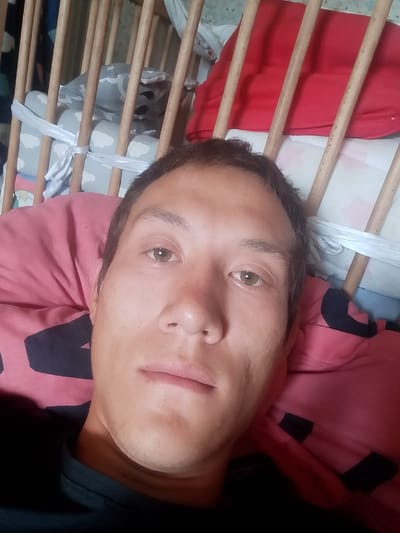 A close-up, selfie-style, horizontal, eye-level shot depicts a young man lying in a crib. The man's face is the primary subject, filling most of the frame. He has light brown skin, short dark brown hair, and light brown eyes. His facial expression is neutral. He is wearing a black t-shirt. The crib features light brown wooden slats, and soft bedding with a pink pillow and a light gray patterned blanket is visible. The background is the interior of the crib. The location is specified as Lobanovo, Kazakhstan.