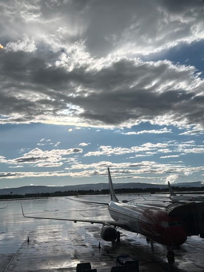 The image shows a wet tarmac at Oslo Airport Gardermoen, Norway. A red and white Norwegian Air Shuttle Boeing 737 aircraft is parked at a gate, connected to a jet bridge. The tarmac is reflecting the cloudy sky, and there are other aircraft visible in the distance. The sky is filled with dramatic, layered clouds, with patches of blue sky and sunlight breaking through. In the background, a tree line and hills can be seen. The overall impression is of an airport scene during or after rain.