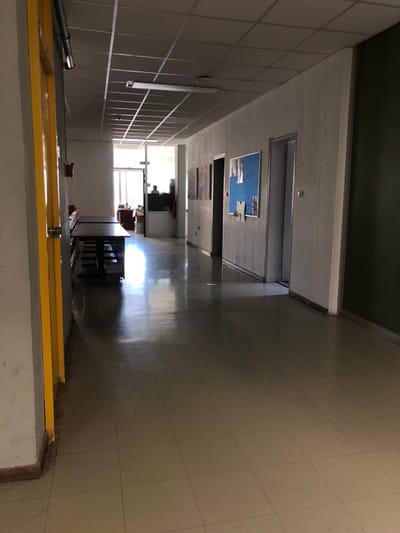 The image shows a hallway in a building located in Agia Paraskevi, Greece, at the coordinates 37.998, 23.82. The hallway is long and narrow, with tiled walls and a tiled floor. The walls are painted white, and the floor is a light gray. There are several doors on either side of the hallway, all closed. One door, at the very end of the hall, is partially open, revealing a room with a window. The hallway is empty, with no people present. There is light coming in from the window at the end of the hall, casting a bright light on the floor. The ceiling has a grid of light brown square panels. A bright yellow door frame extends from the bottom of the image to the left, and a concrete wall forms the right edge of the image. There is a shadow on the wall in the lower left corner. The image is taken from a low angle, looking down the hallway.
