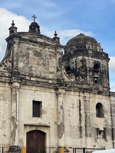 The image depicts the exterior of the Basílica Catedral de la Natividad de María in León, Nicaragua. The cathedral's facade is aged and weathered, with peeling paint and visible signs of deterioration. The architecture features a central dome, bell tower, ornate carvings, and arched windows. The sky above is a mix of blue and white clouds. A cross is visible atop one of the cathedral's spires. The foreground shows a black metal fence, and a portion of a yellow structure is visible to the left. Several birds are perched on the building's ledges and cornices.