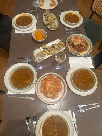 This image depicts an indoor dining scene, presumably a dinner or shared meal, taking place in Rueil-Malmaison, France. The view is from a high angle, looking down onto a dark brown or grey rectangular dining table set for multiple individuals. No people are visible in the frame.
The table is extensively covered with various food items, beverages, and dinnerware. Four main place settings are discernible, each featuring a white bowl containing a reddish-brown stew-like dish with visible legumes and meat chunks, placed atop a larger white plate and a white paper napkin. Cutlery, including a spoon, fork, and knife, is positioned beside each setting. One napkin bears the red text "Semorodes".
Food items present include:
- Four bowls of stew.
- A light green plate holding a round, flatbread, partially consumed.
- A beige plate with an intricately shaped bread, possibly a traditional loaf, topped with black seeds.
- Two elongated white platters, each holding multiple triangular fried or baked pastries (possibly samosas or briouats) garnished with lime wedges.
- A white plate with several rectangular flat pastries, possibly msemen.
- A small white bowl containing orange nacho-style corn chips.
Beverages consist of multiple clear stemmed wine glasses, some containing white wine, and a tall clear water glass. Portions of dining chairs, one with a rattan-style back and a green upholstered seat, are visible at the edges of the frame. A light-colored wooden floor is discernible in the background. The arrangement suggests a meal is either in progress or prepared for consumption.