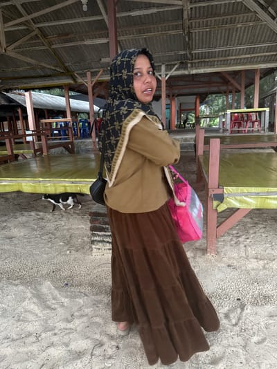 A full-shot, eye-level, outdoor medium-light natural light portrait shows a young Indonesian woman, wearing a black and gold patterned hijab and a brown tiered skirt, standing on a sandy surface under a covered wooden structure. She faces the camera with her head turned to her right, her mouth slightly agape. A brown jacket is worn over a light-colored garment, and a black shoulder bag is slung across her right shoulder. A bright pink tote bag hangs from her right hand. Behind her, a black and white cat walks away from the camera towards the left. The background reveals a sandy area with scattered debris, a brick pillar on the right, and a raised wooden platform with yellow coverings. Further back, a wooden structure with a corrugated roof and exposed beams is visible, as well as rows of red chairs and tables. The location is identified as Meunasahbale, Indonesia.