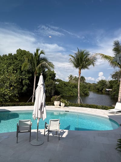 This image depicts a backyard swimming pool area in East Naples, Florida, United States of America. The scene includes an oval-shaped swimming pool filled with clear blue water, surrounded by a light-colored paved patio. Two white chairs with mesh seats and a closed white umbrella on a stand are positioned on the patio near the pool. In the background, lush green trees and palm trees line a dark body of water, likely a canal or estuary. A small rustic structure with a roof is visible on the opposite bank of the water. The sky is a bright blue with scattered white clouds. There are no people visible in the image.