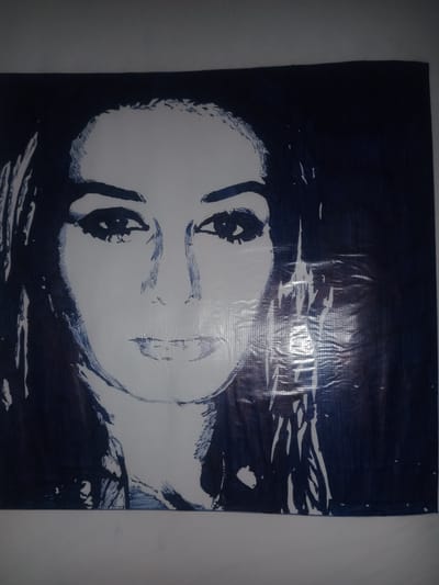 This is a close-up, portrait-style artwork of a woman's face rendered in a stark, high-contrast style with dark blue ink on a white surface. The artwork appears to be on a wrinkled or textured material, possibly plastic sheeting, which creates a subtle sheen and visible horizontal lines across the image. The woman's features are outlined and shaded with bold strokes, emphasizing her eyes, eyebrows, nose, and mouth. Her expression is neutral or perhaps slightly somber. Dark, stylized hair frames her face. The background is a solid dark blue, creating a strong silhouette effect. The overall aesthetic is graphic and somewhat dramatic, reminiscent of a stencil or a graphic novel illustration. There is no visible text within the artwork. The setting is not explicitly depicted, but the image focuses solely on the portrait.