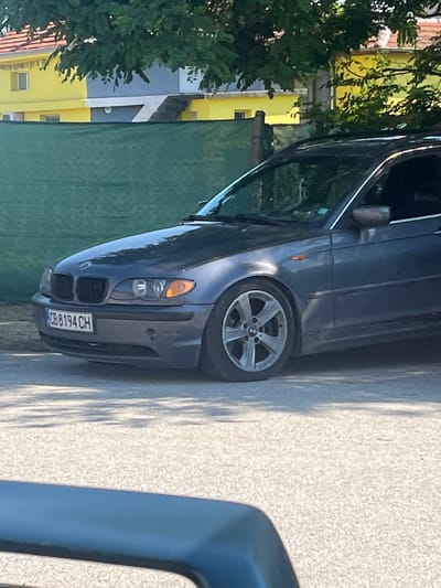 A gray BMW sedan is parked on a paved surface adjacent to a green-painted wall and a tree in Trilistnik, Bulgaria. The front of the car, with license plate "CB 8194 CH", is visible. A portion of a building painted in yellow and gray can be seen in the background behind the tree.